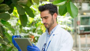 Man holding a lab flask while using a tablet in a greenhouse